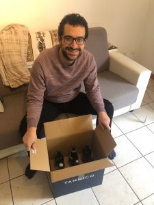 Luca sits on a cream sofa and in front of him is a box with six bottles of wine in it