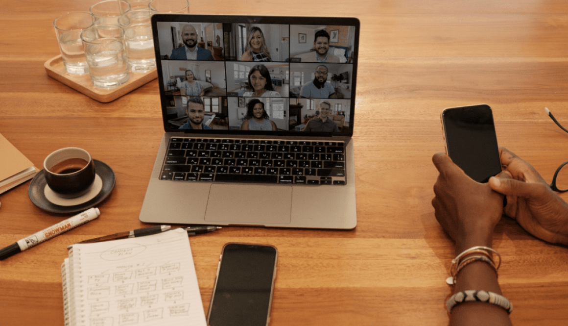 Laptop on the table. Screen shows multiple people attending an online event. Items such as a coffee cup, notebook and pens are scattered around the laptop.