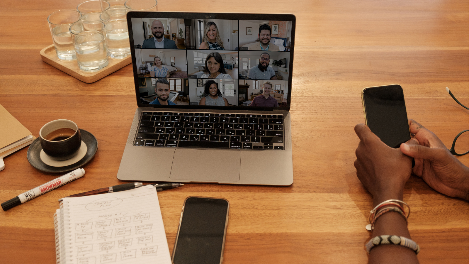 Laptop on the table. Screen shows multiple people attending an online event. Items such as a coffee cup, notebook and pens are scattered around the laptop.